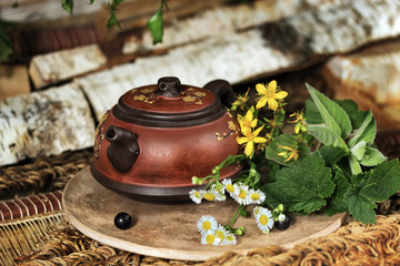 Obraz na płótnie Canvas Clay teapot with herbal tea is in the sauna on a mat. Near wild flowers, firewood and birch brooms. Horizontal