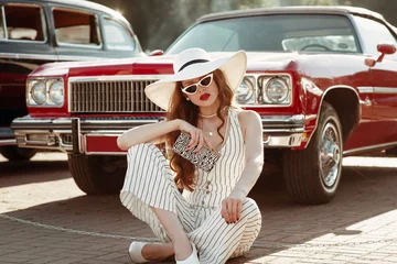 Straßenmodeporträt in voller Länge der jungen eleganten Luxusdame mit weißer Sonnenbrille, Hut mit breiter Krempe, gestreiftem Leinenoverall, Zebra-Geldbörse mit Animal-Print in der Hand, posiert in der Nähe eines roten Autos. Platz kopieren © Victoria Fox