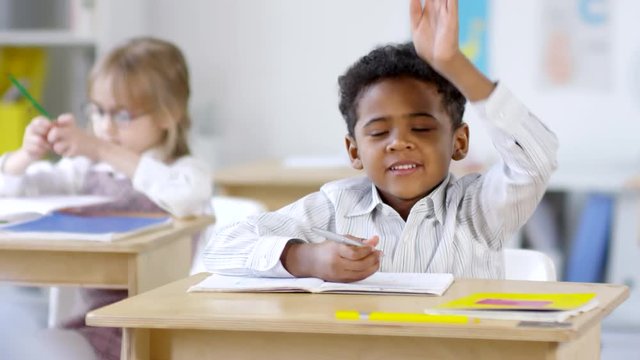 Medium shot of cute little African-American schoolboy sitting at his desk and writing in exercise book, then raising hand and answering question in class