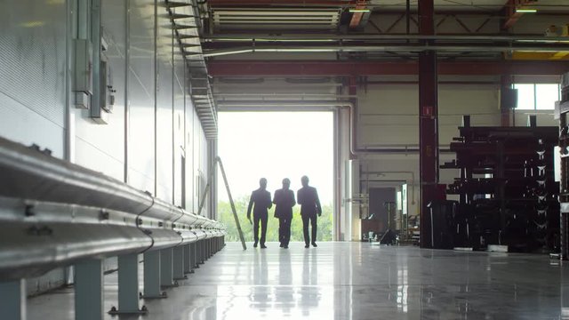 Panning long shot of three directors wearing hardhats arriving at industrial plant, filmed against bright sunlit entrance, walking towards camera engaged in discussion