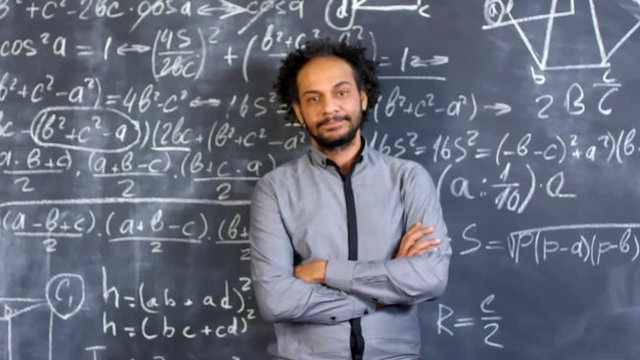 Dolly-in portrait shot of smart male mathematician with serious expression on his face standing with his arms crossed before blackboard with complex calculus formulas