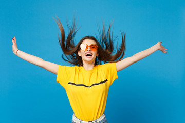 Fototapeta na wymiar Portrait of laughing young woman in vivid casual clothes heart eyeglasses with flowing hair spreading hands isolated on blue wall background in studio. People lifestyle concept. Mock up copy space.