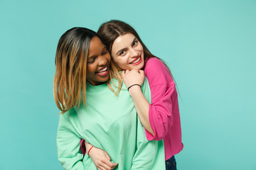 Two young women friends european and african american in pink green clothes standing posing isolated on blue turquoise wall background, studio portrait. People lifestyle concept. Mock up copy space.