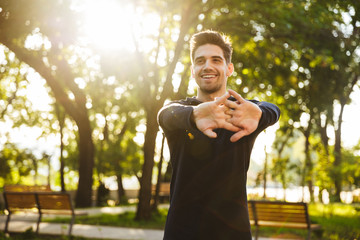 Cheerful young sports fitness man standing in green park nature make stretching exercises for arms.