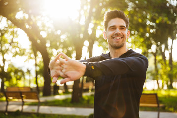 Cheerful young sports fitness man standing in green park nature make stretching exercises for arms.