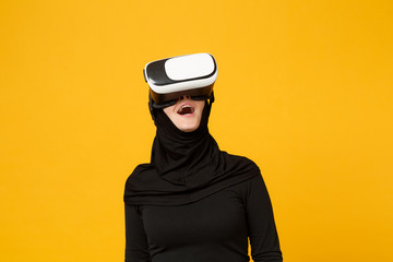 Young arabian muslim woman in hijab black clothes watching in headset of vr virtual reality isolated on yellow wall background, studio portrait. People religious lifestyle concept. Mock up copy space.