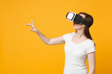 Young woman watching in headset of VR, touch something like push click on button pointing at floating virtual screen isolated on yellow orange background. People lifestyle concept. Mock up copy space.