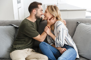 Smiling young loving couple indoors at home sitting on sofa hugging kissing.