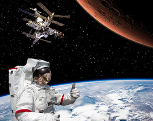 Astronaut shows the thumbs-up about flying to Mars. Space station on the backdrop. The elements of this image furnished by NASA.