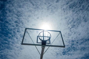 Basketball basket on a background of blue sky with light clouds. The sun is right above the basket like a ball. Place for text. The concept of summer sports, summer hobby.
