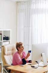 Pretty aged woman sitting at computer table and using credit card to pay her bills online