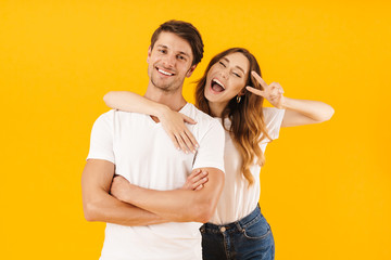 Naklejka premium Portrait of joyous couple in basic t-shirts standing together while woman hugging man and showing peace sign at camera