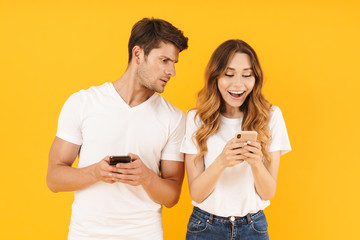 Photo of young couple man and woman in basic t-shirts holding and peeking at cell phones