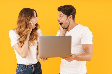 Portrait of excited couple man and woman in basic t-shirts rejoicing while standing together with silver laptop