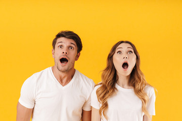 Portrait of shocked couple man and woman in basic t-shirts wondering while looking upward with open mouthes at copyspace