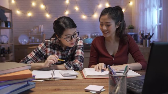 University female students sitting together at table at midnight. two young people working with textbook and doing project for final exam in home kitchen at night. girls sharing ideas and discussing.