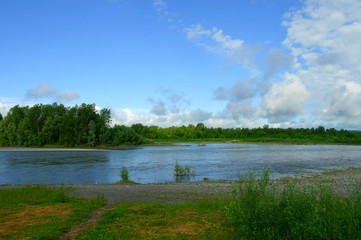 Landscape, blue river flows. Along the banks is bright green grass and forest. Above them is a blue sky with white clouds.