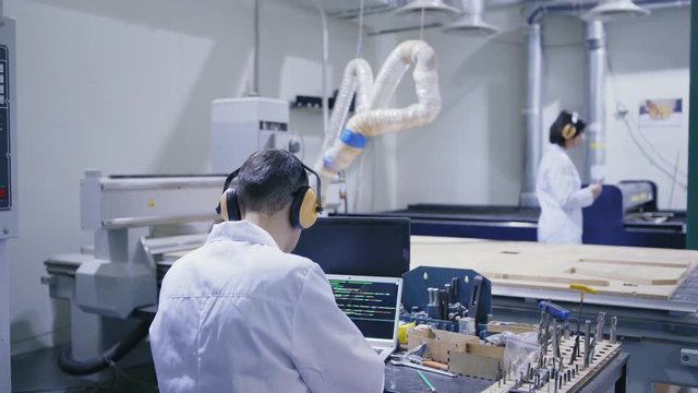 Engineer in white coat, protective eyewear and ear muffs working on laptop in laboratory, female technician showing him plywood detail produced by cnc machine