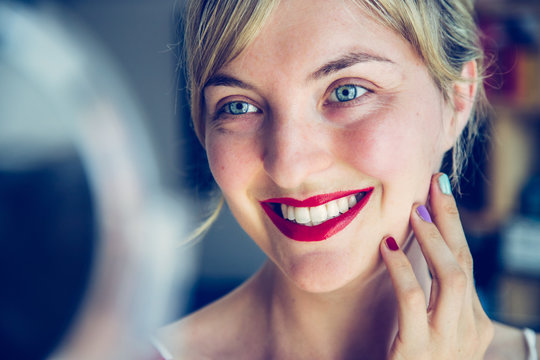 Attractive red lip stick and healthy white teeth: Blonde young woman with colorful nail polish is looking herself in the mirror.