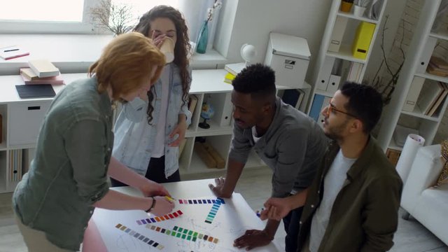 High-angle shot of four multicultural millennial graphic designers standing together in studio around table, discussing layout of new project and choosing colours from selection of palettes