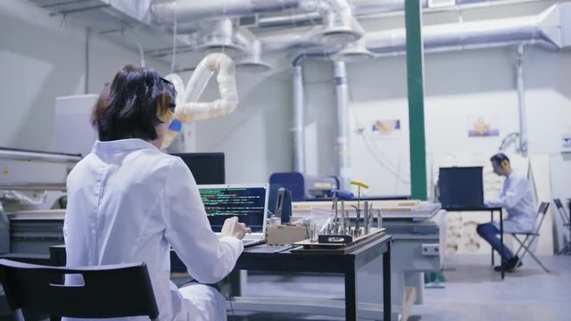 Tracking right back view shot of female engineer in white coat and safety ear muffs working on laptop computer in cnc laboratory with laser engraving machines
