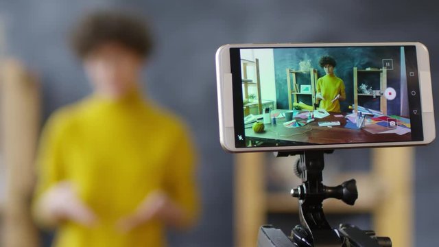 Tracking shot with rack focus of young female video blogger in glasses talking to camera when recording vlog on smartphone fixed on tripod