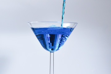 Cocktail with blue liquid in glass. Glass with blue water pouring with liquid with splashes and drops. Martini glass filling with alcohol with splashes on white background. Refreshing drink concept.