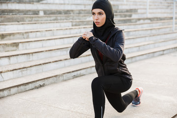 Muslim serious sports fitness woman dressed in hijab posing make sport stretching exercises outdoors at the street with steps on background.