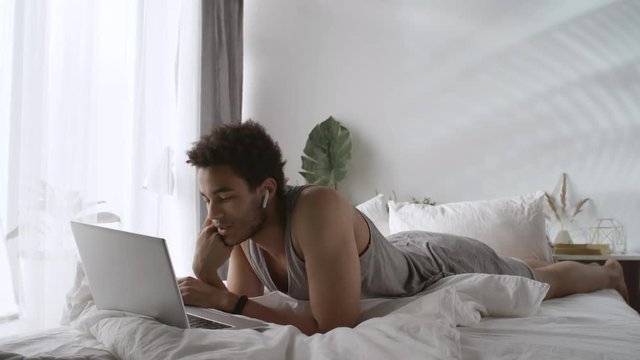 Young black man lying on bed after waking up early in the morning and using laptop computer and wireless earphones