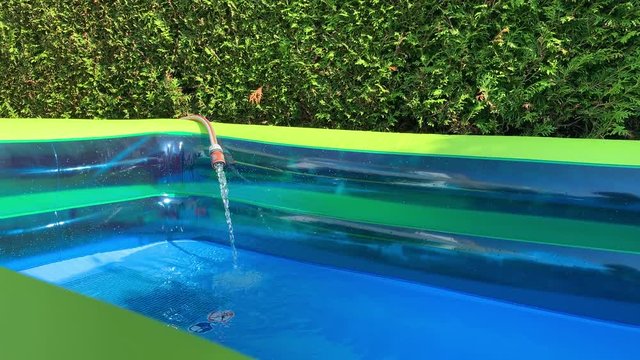 [Slide] Inflatable outdoor pool is filled with water. Nozzle and hose filling plastic pool with water. View from pool edge.