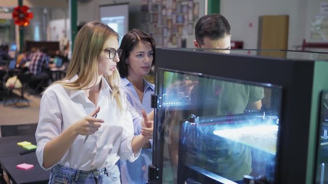 Team of three creative engineers examining models being printed in 3d printing machine in laboratory, medium shot