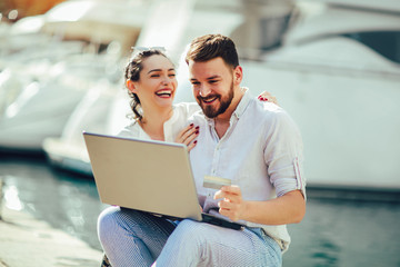 Beautiful romantic couple using laptop. Watching pictures on the laptop while traveling, by the harbor of a touristic sea resort with boats on background.