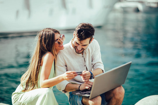 Beautiful romantic couple using laptop. Watching pictures on the laptop while traveling, by the harbor of a touristic sea resort with boats on background.