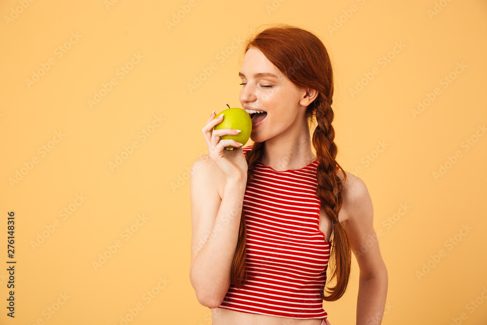 Sticker happy young beautiful redhead woman posing isolated over yellow background eat apple.