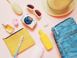 Summer trip stuff, girly travel items on a pink background. Cosmetic bag, perfume, yellow sunscreen bottle, shampoo, toothbrush, hair brush, sunglasses, camera, sun hat and jeans. Flat lay photo