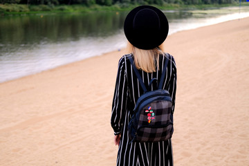 Fototapeta premium Blonde girl in black hat with backpack walking on the street. City traveling alone concept