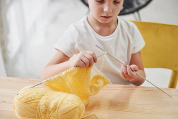 Doing homework. Kid is knitting. Cute little girl sitting near the wooden table is learning some new stuff