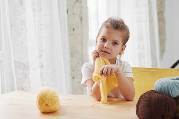 Fototapeta premium Time for the rest. Kid is knitting at home. Cute little girl sitting near the wooden table is learning some new stuff