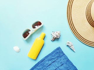 Flat lay photo beach essentials. Sunglasses, towel, yellow sunscreen bottle, sun hat and seashells on a blue background