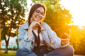 Fototapeta premium Smiling cheery cute young student girl wearing eyeglasses sitting on bench outdoors in nature park with beautiful sunlight using mobile phone.