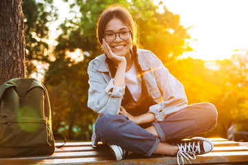 Happy cute young student girl wearing eyeglasses sitting on bench outdoors in nature park with beautiful sunlight.