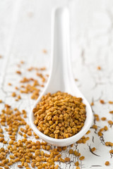 Dried, raw fenugreek seed in white spoon close up on white rustic wooden table background
