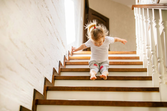 Baby blonde girl in white t-shirt at bottom of stairs indoors, looking at camera and smiling