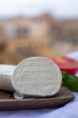 Italian mozzarella soft cheese ball served with basil and tomato on wooden plank with yellow houses in old Italian town on background