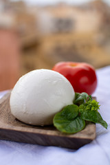 Italian mozzarella soft cheese ball served with basil and tomato on wooden plank with yellow houses in old Italian town on background