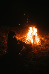 Woman sitting and getting warm near the bonfire in the night forest.