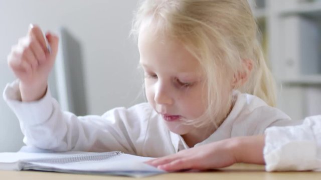 Handheld shot of adorable little girl sitting at her desk and writing in notebook, then talking to someone off camera and smiling