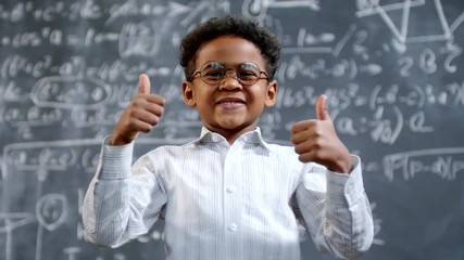 Portrait shot of happy little African-American schoolboy in glasses standing before blackboard with complex calculus formula and showing thumbs up