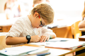 Blonde boy with big black glasses sitting in classroom, studing, smiling. Education on elementary...