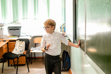 Fototapeta na wymiar Little boy with big black glasses and white shirt standing near school blackboard with a piece of chalk making smart thinking face